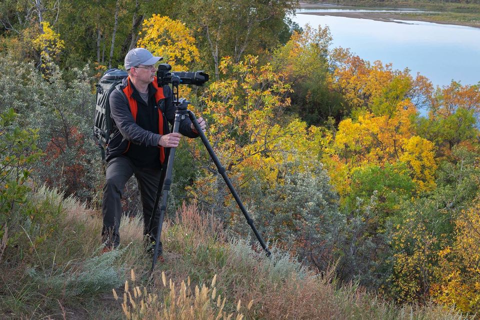 Photographer with camera on tripod standing on hill near riverbank.