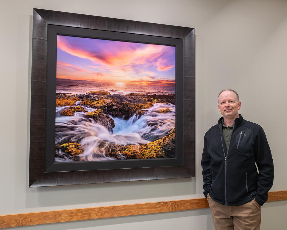 Man posing beside framed print of coastal sunset hanging on wall.