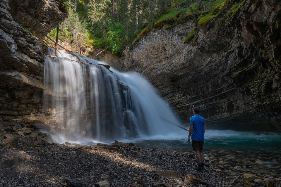 Man standing with back to camera in front of waterfall.
