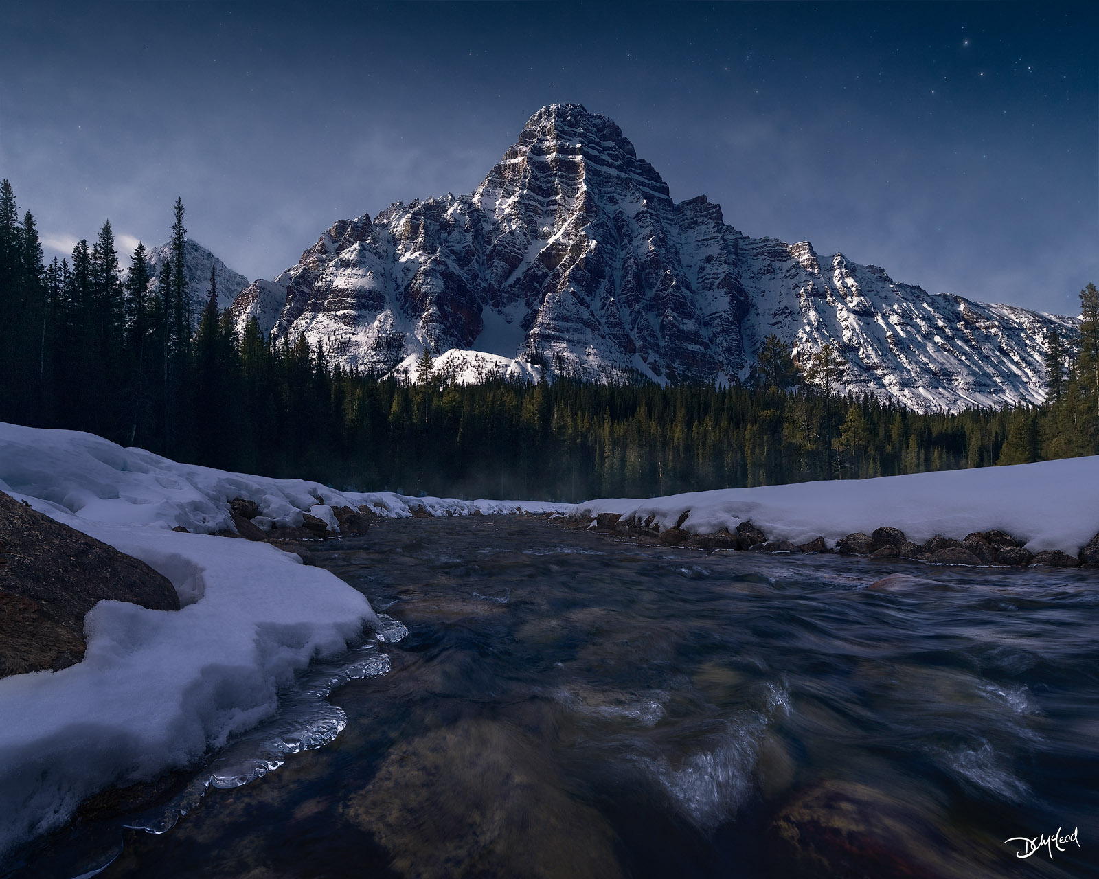 Mount Chephren under moonlight in the winter with snow and ice formations along the river in Banff National Park.
