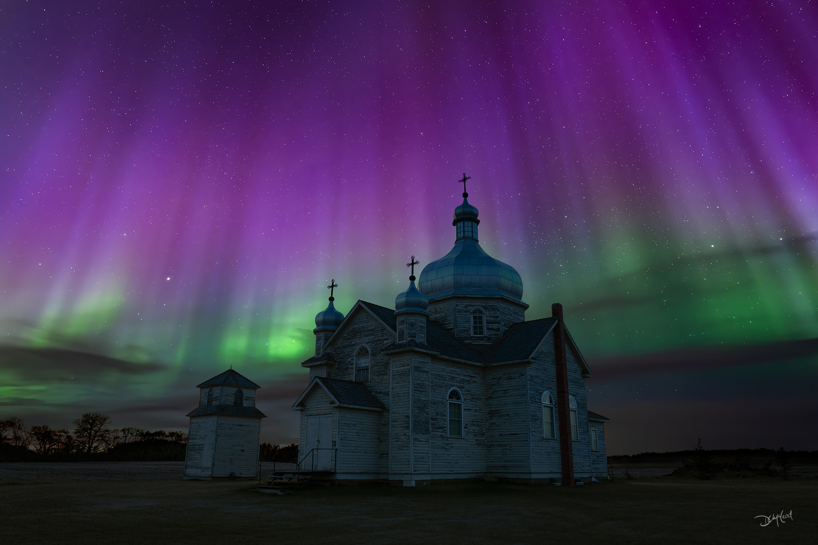 Ss. Peter and Paul Ukrainian Catholic Church (Bodnari Church) illuminated at night beneath colourful pink and green aurora borealis.