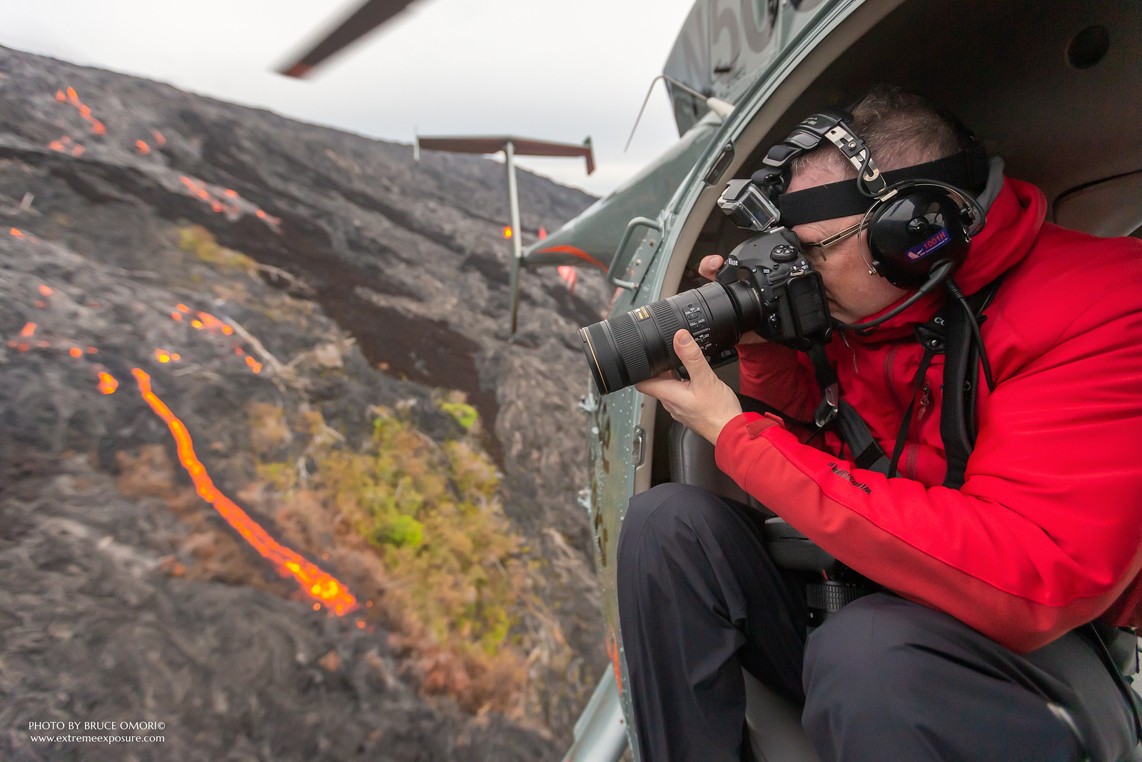 A photographer hovering in a doors-off helicopter shooting a lava field in Hawaii.